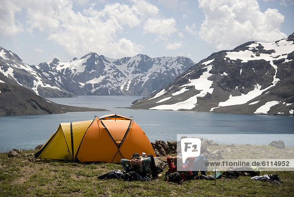 Two men rest next to their orange tent enjoying the dramatic views over a high alpine lake.