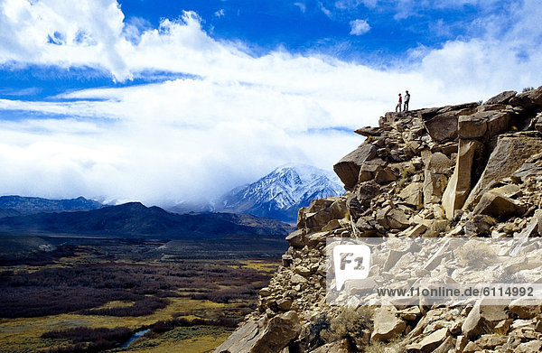 A couple stands on top of a lava butte overlooking a grassy valley with a snow capped peak in the background.