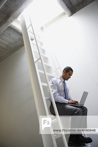 Businessman sitting on ladder with laptop