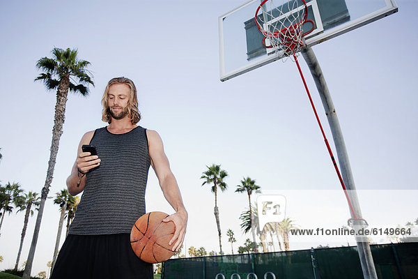 Caucasian man holding basketball and text messaging on cell phone