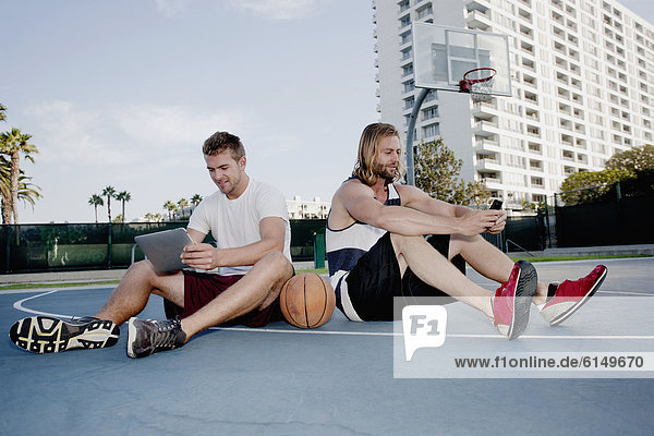 Caucasian men using cell phone and digital tablet on basketball court