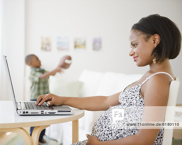Pregnant Black mother using laptop while son plays with football