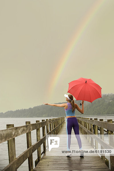 Caucasian woman in sportswear with red umbrella on pier catching rainbow