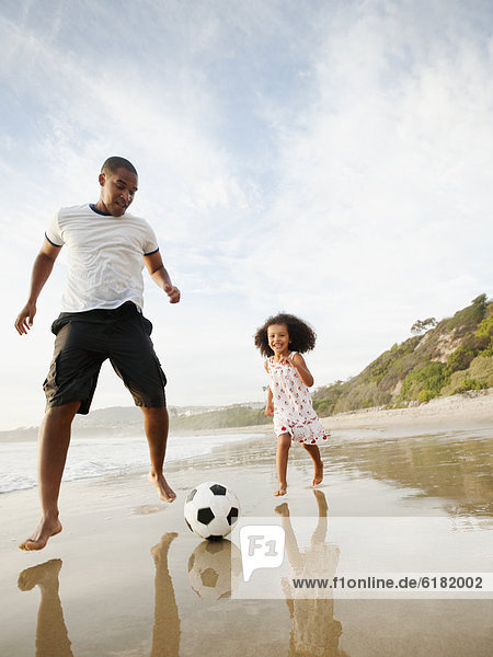 Black father kicking soccer ball with daughter on beach