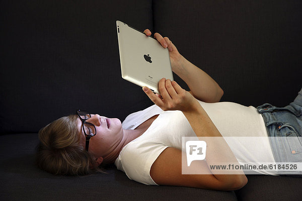 Junge Frau liegt auf einem Sofa und surft mit einem iPad Tablet-Computer drahtlos im Internet