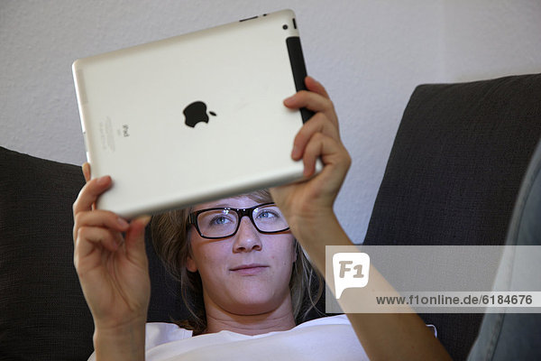 Junge Frau liegt auf einem Sofa und surft mit einem iPad Tablet-Computer drahtlos im Internet