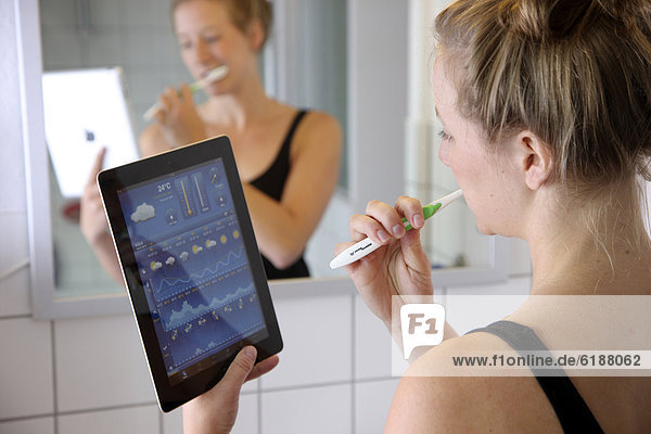 Junge Frau liest morgens beim Zähne putzen im Badezimmer Nachrichten auf einem iPad Tablet-Computer per drahtlosem Internetzugang