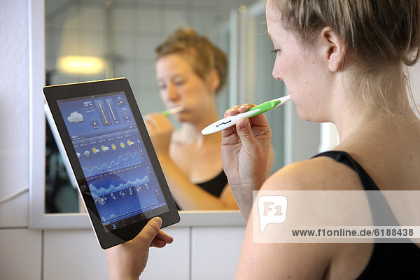Junge Frau liest morgens beim Zähne putzen im Badezimmer Nachrichten auf einem iPad  Tablet-Computer  per drahtlosem Internetzugang