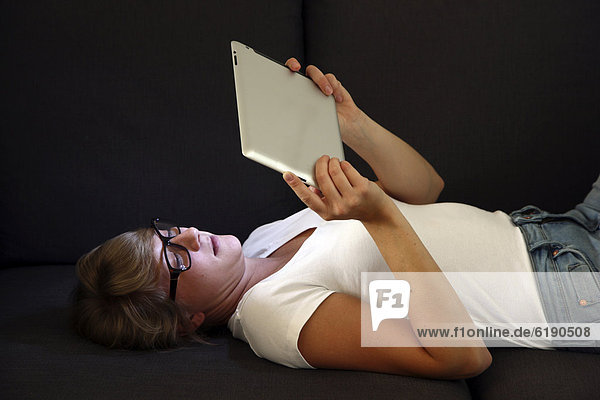 Junge Frau liegt auf Sofa und surft drahtlos im Internet mit einem iPad Tablet-Computer