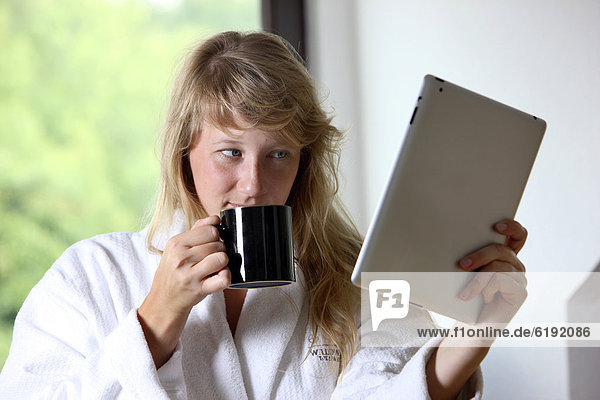 Junge Frau im Bademantel morgens mit einer Tasse Kaffee  liest mit einem iPad  Tablet-Computer in einer Online-Zeitung
