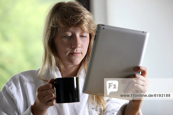 Junge Frau im Bademantel morgens mit einer Tasse Kaffee  liest mit einem iPad  Tablet-Computer in einer Online-Zeitung