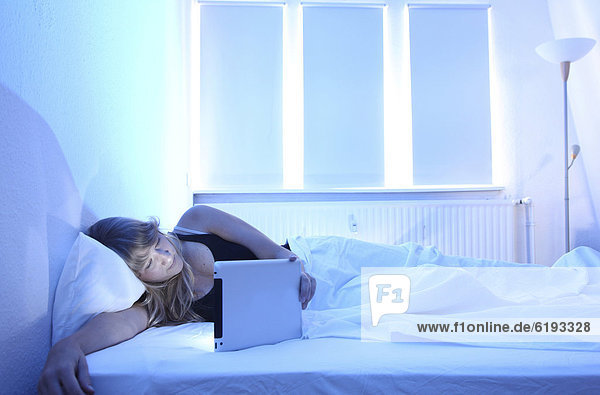 Junge Frau im Bett surft mit einem iPad Tablet-Computer per Drahtlosverbindung im Internet