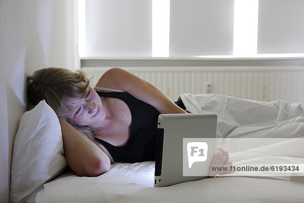 Junge Frau im Bett surft mit einem iPad Tablet-Computer per Drahtlosverbindung im Internet