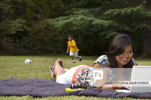 Asian mother using laptop in park with son in background