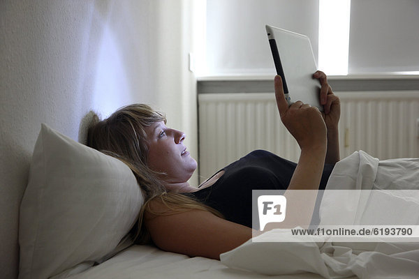Junge Frau im Bett surft mit einem iPad Tablet-Computer per Drahtlosverbindung im Internet