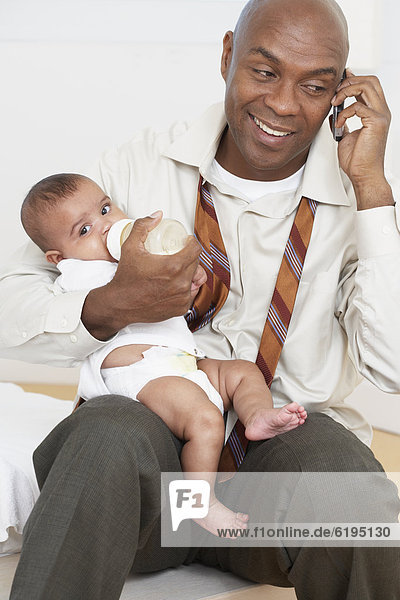 African father feeding baby daughter and talking on cell phone