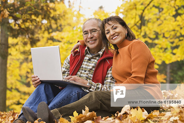 Hispanic couple using laptop sitting in autumn leaves