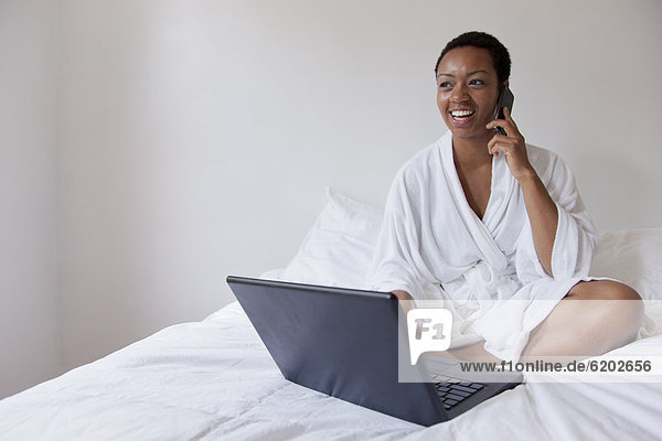 African American woman using laptop and cell phone in bed