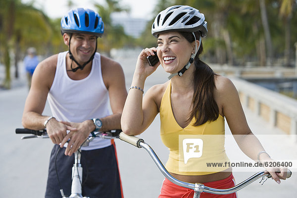 Hispanic woman on bicycle talking on cell phone while boyfriend waits