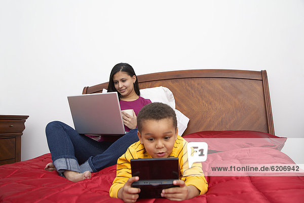 Dominican woman using laptop on bed  son playing video game