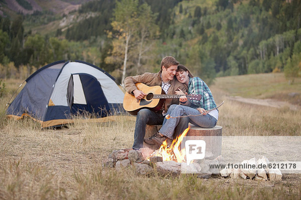Caucasian man playing guitar for wife near campfire