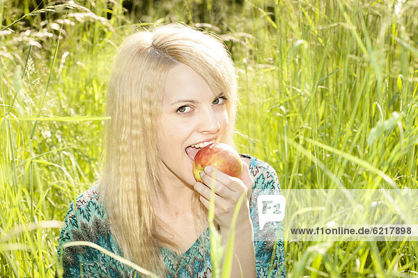 Lächelnde junge Frau sitzt im hohen Gras und isst einen Apfel