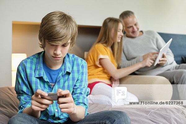 Teenager-Junge mit Handy und Vater und Schwester mit digitalem Tablett im Hintergrund