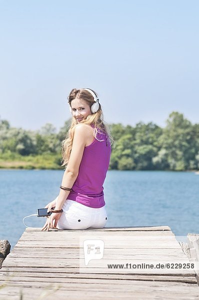 Blonde junge Frau mit Kopfhörern auf einem Steg am See