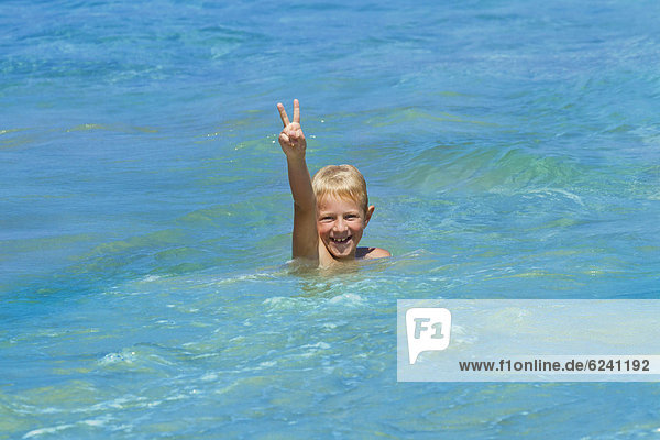 Ein Junge  8 Jahre  badet lachend im Meer  macht V-Zeichen