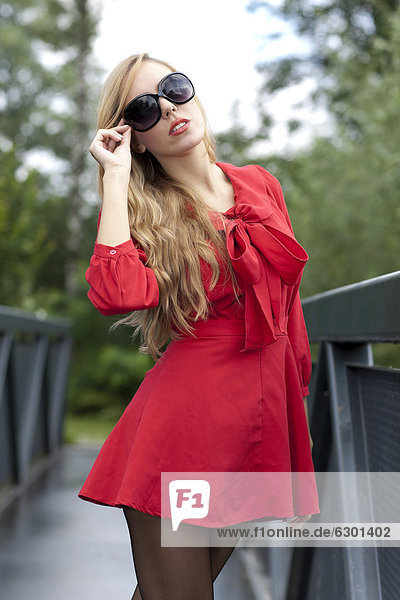 Young woman in a short red dress and sunglasses posing on a bridge