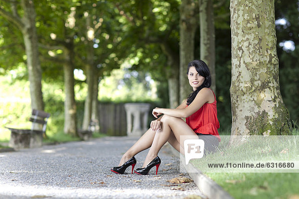 Junge Frau mit rotem Top  schwarzen Hotpants und hochhackigen Schuhen posiert sitzend vor Baum