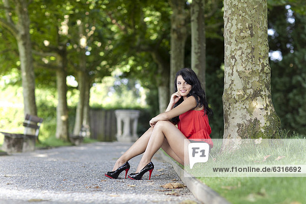 Junge Frau mit rotem Top  schwarzen Hotpants und hochhackigen Schuhen posiert sitzend vor Baum