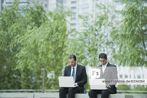Geschäftsleute sitzen Seite an Seite und benutzen Laptops im Freien.