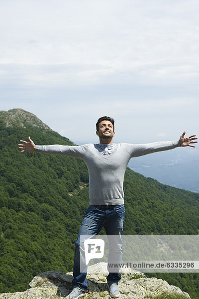 Mid-adult man standing at edge of rock with arms outstretched  mountainscape in background