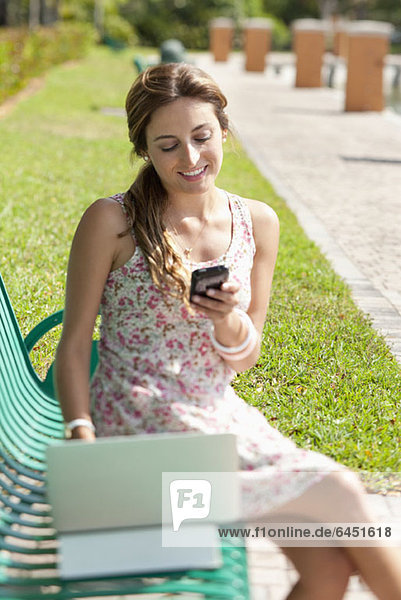Eine Frau sitzt auf einer Parkbank mit einem Laptop und einem Handy.