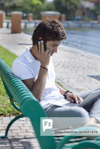 Ein Mann  der auf einer Parkbank sitzt  auf einem Handy spricht und einen Laptop benutzt.