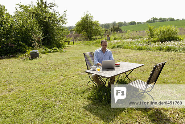 Ein Mann  der an einem Tisch in seinem Garten sitzt  frühstückt und einen Laptop benutzt.