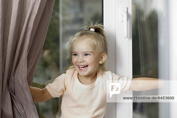 Ein junges lachendes Mädchen schiebt Fenstervorhänge auseinander.