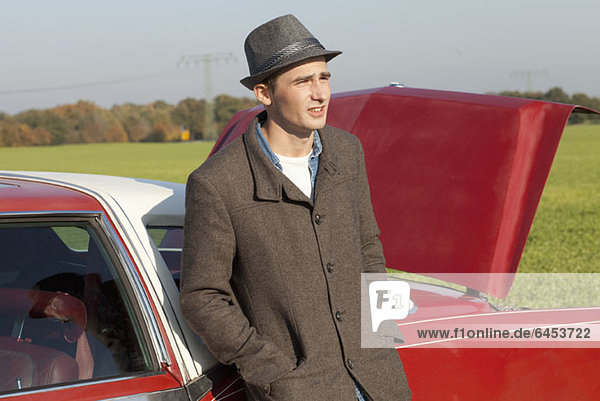 A young rockabilly man leaning against a vintage car in the country