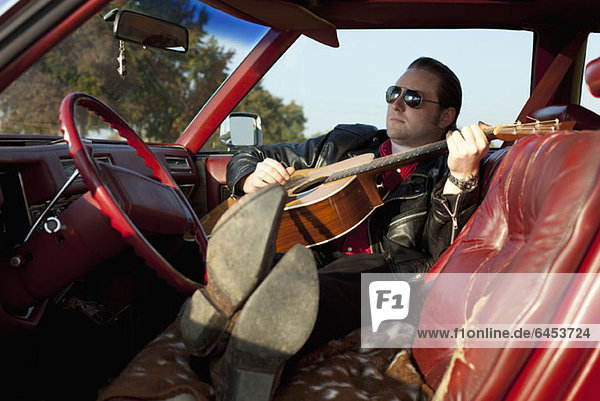 A rockabilly guy playing an acoustic guitar while sitting in his vintage car
