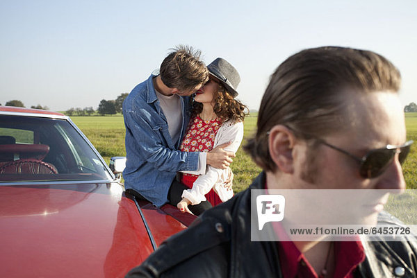A rockabilly couple kissing while leaning on a vintage car  man in foreground