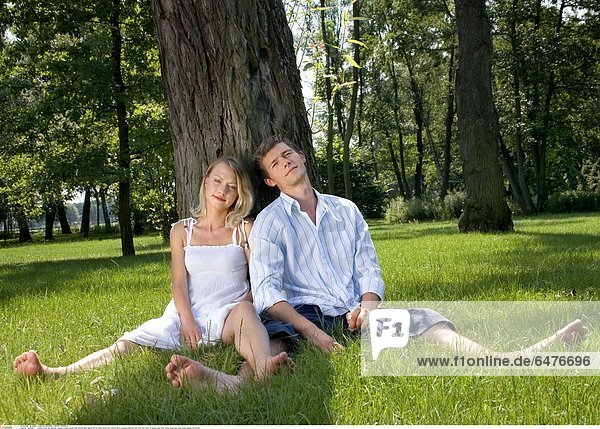 1224889 outdoor park day summer couple young woman man blonde dark haired 25-30 white dress shirt shorts short trousers barefot foot feet rest relax sit grass tree trunk resist close eye eyes smile smiling horizontal