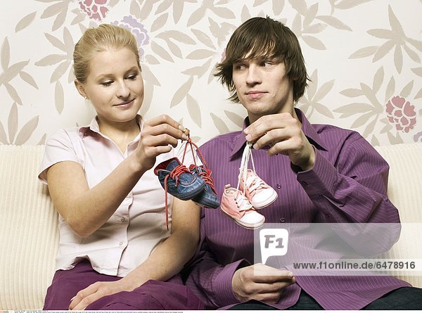 1225354 indoor flat room wall pattern patterns flower flowers people woman couple 25-30 blonde man young 30-35 dark haired blouse violet pink blue blouses sofa couch sit smile smiling hold shoe shoes close up symbole pregnancy embrace show astonishment surprise mime message horizontal