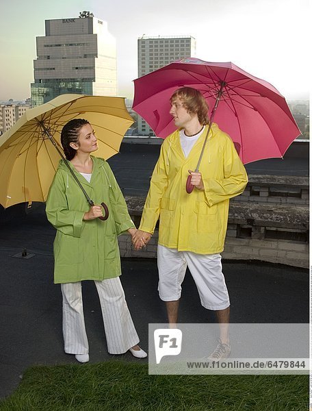 1229545 outdor roof city day summer spring people couple young man woman fair haired boy 20-25 girl woman brunette pigtail pigtails stand white trousers short shorts yellow green raincoat raincoats hold hand hands umbrella umbrellas rain raining vertical