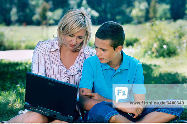 1228640 outdoor day summer people woman 35-40 mature blonde blouse white stripe stripes striped lie laptop internet personal computer notebook communication rest relax field sit smile smiling horizontal write show vacation boy son 10-15 dark haired hair blouse blue trousers jean jeans