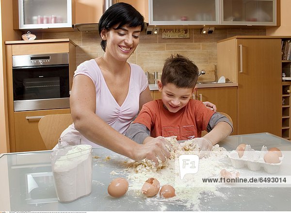 1235617 indoor flat kitchen people child boy 5-10 fair haired blouse orange table egg eggs glass water make cake flour break close up smile smiling mother woman mature 30-35 brunette blouses pink horizontal