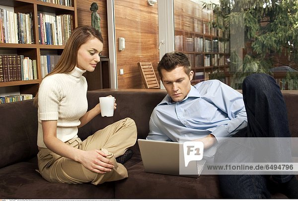 1237082 indoor flat room people couple young woman girl 20-25 brunette long hair sit couch sofa man dark haired 25-30 jean jeans trousers shirt blue jersey white beige cross leg legs hold mug drink laptop notebook personal computer work internet communication smile smiling close up horizontal