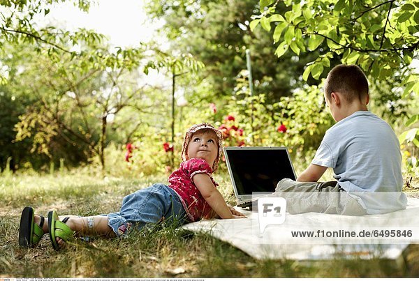 1243628 outdoor day summer people child boy 5-10 fair haired blouse white trousers grey personal computer laptop internet communication sit blanket rest relax read write girl 0-5 blonde cap pink blouse pattern patterns trousers jean jeans sandal sandals sister brother siblings horizontal lie