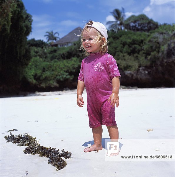 Young Blond Child Standing On Beach By Seaweed