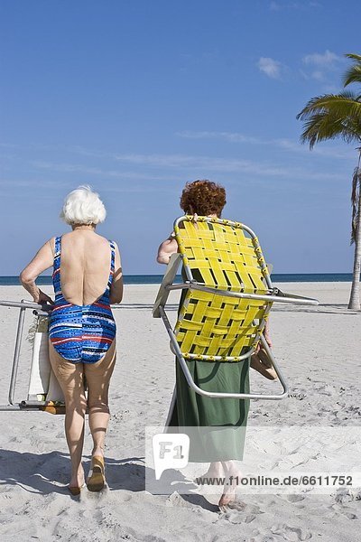 Senior women carrying deck chairs walking on beach
